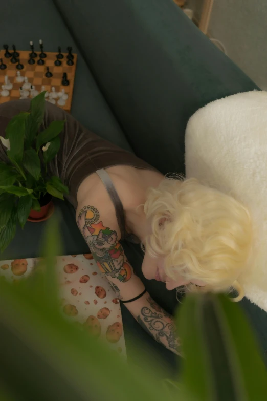 a woman laying on a couch next to a potted plant, a tattoo, inspired by Elsa Bleda, reddit, low quality footage, albino hair, high angle close up shot, low quality photo