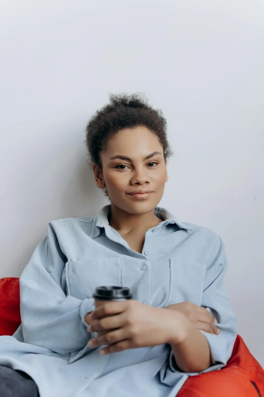 a woman sitting on a red couch holding a cell phone, light-brown skin, on a gray background, with a white mug, hair styled in a bun
