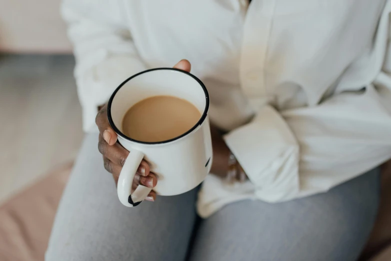 a woman sitting on a bed holding a cup of coffee, by Emma Andijewska, trending on unsplash, minimalism, holding a boba milky oolong tea, sitting on a sofa, sitting in office, wearing a white sweater