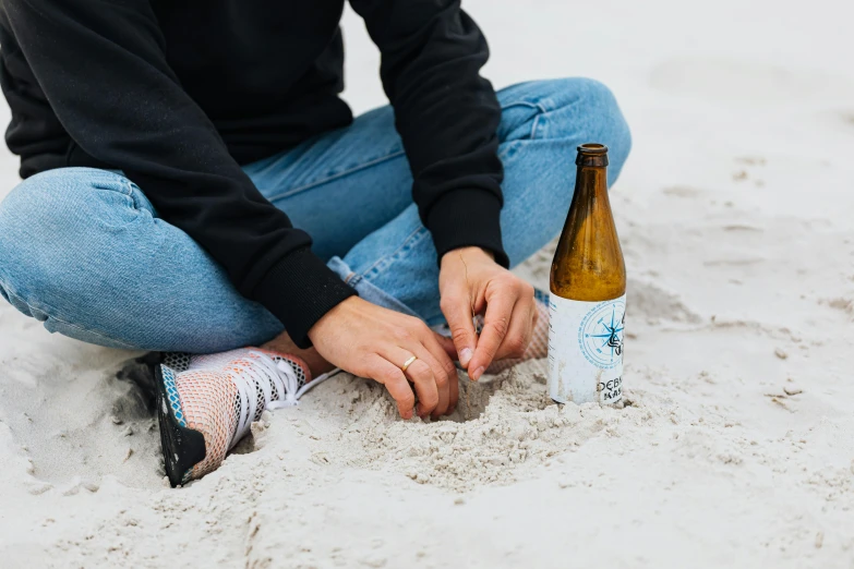 a woman sitting in the sand with a bottle of beer, by Tuvia Beeri, unsplash contest winner, new england ipa, intricate detailing, manly, detailed white