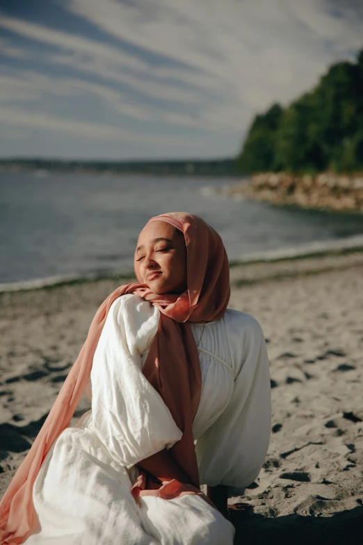 a woman sitting on top of a sandy beach, a picture, inspired by Nazmi Ziya Güran, pexels contest winner, hurufiyya, white hijab, seattle, in shades of peach, [ cinematic