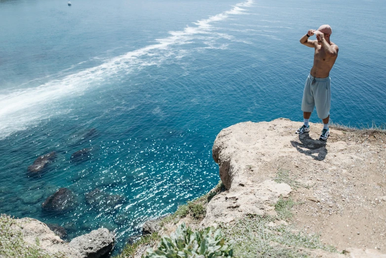 a man standing on top of a cliff next to the ocean, cyprus, white shorts and hiking boots, set photo, illustration »