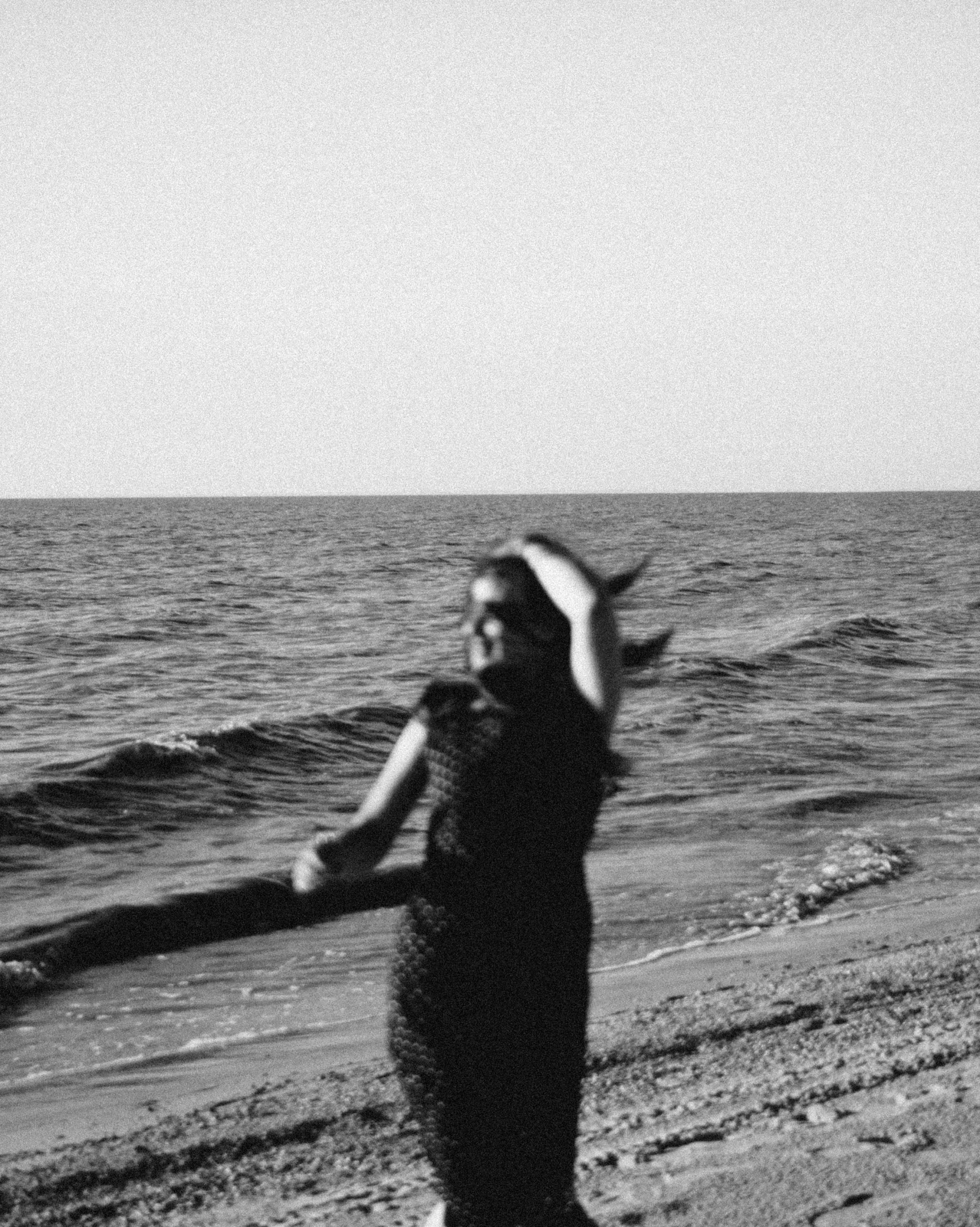 a woman standing on top of a beach next to the ocean, a black and white photo, by Lucia Peka, hair waving in the wind, slightly pixelated, photo of the girl, shrugging arms