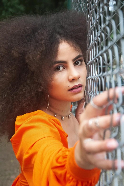 a woman leaning against a chain link fence, an album cover, inspired by Elsa Bleda, pexels contest winner, curly afro, orange, young middle eastern woman, behind bars