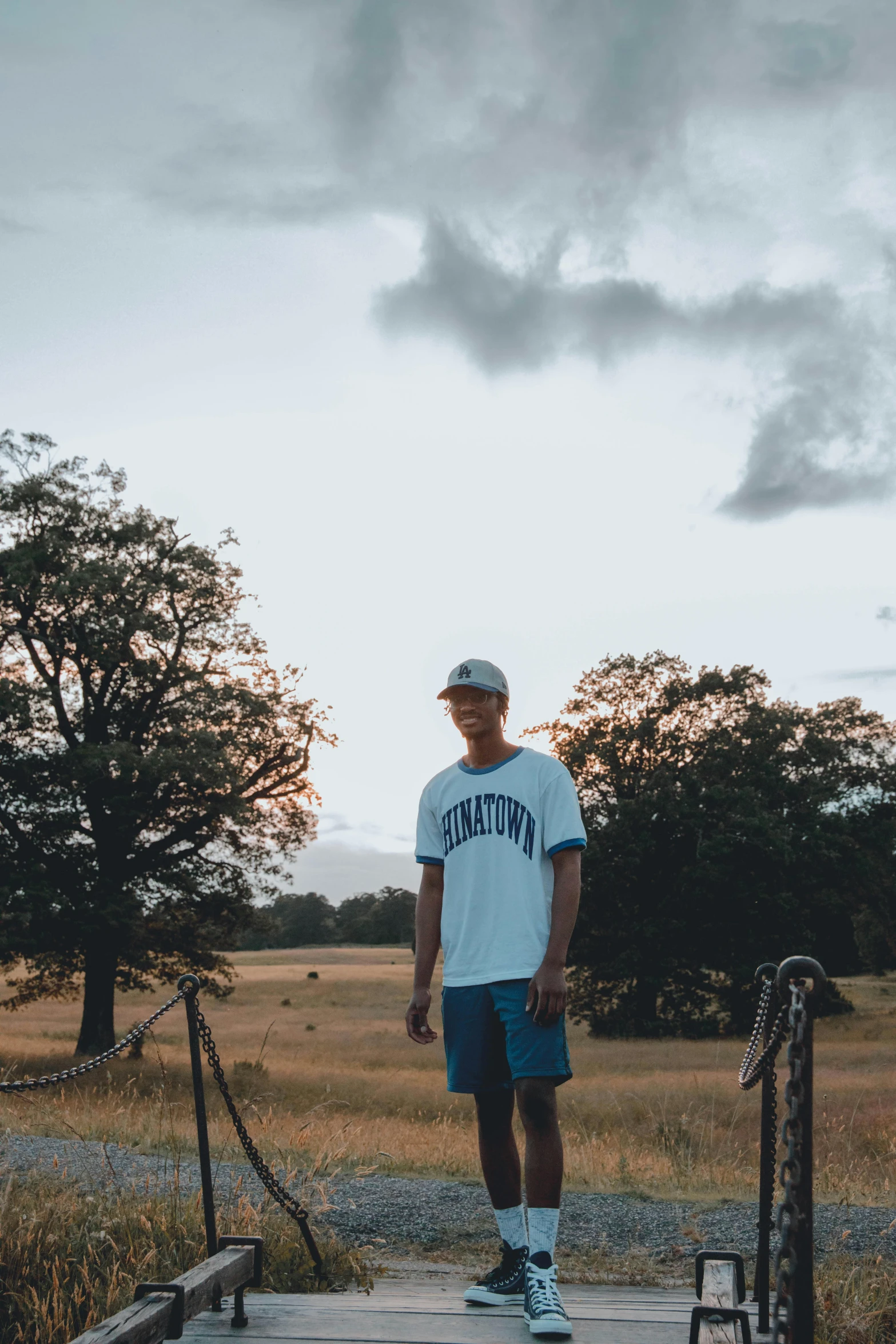 a man standing on a wooden platform in a field, by Andrew Stevovich, wearing shorts and t shirt, trending on vsco, baggy clothing and hat, avatar image