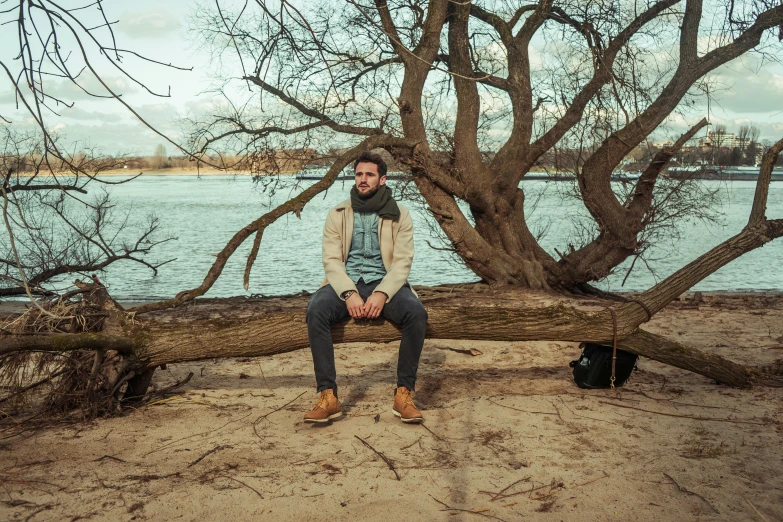 a man sitting on a tree branch next to a body of water, an album cover, inspired by Henry Heerup, pexels contest winner, very attractive man with beard, casual pose, matej ‘retro’ jan, bench