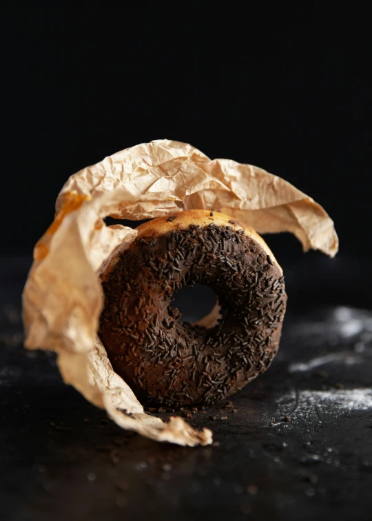 a donut that is sitting on a table, carbon black and antique gold, parchment paper, fully chocolate, alessio albi