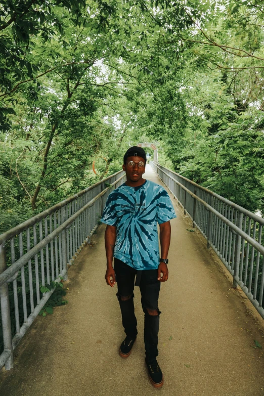 a man standing on a bridge with trees in the background, an album cover, unsplash, wearing a tie-dye t-shirt, ( ( dark skin ) ), looking straight to camera, in style of tyler mitchell