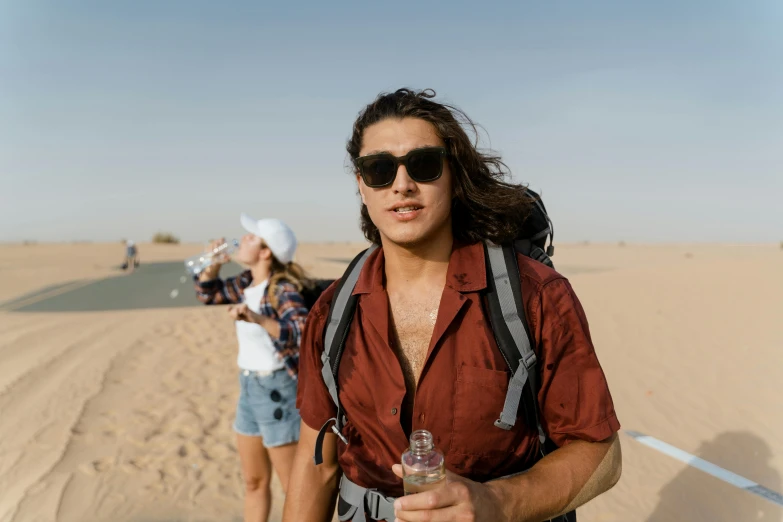a man standing on top of a sandy beach, trending on pexels, happening, avan jogia angel, carrying a bottle of perfume, in the australian desert, a group of people