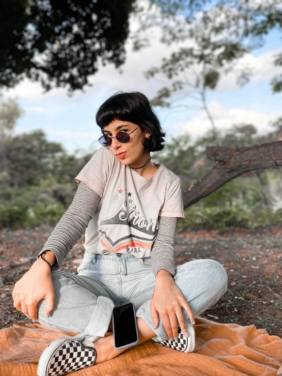 a woman sitting on a blanket with a cell phone, a picture, by Robbie Trevino, trending on pexels, graphic tees, wear ray - ban glass, vintage muted colors, panoramic view of girl