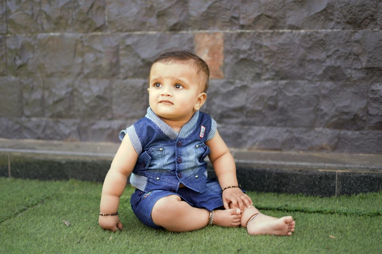a baby sitting on the grass in front of a stone wall, by Saurabh Jethani, pexels, hyperrealism, wearing blue jean overalls, full body image, with accurate face, ( ultra realistic