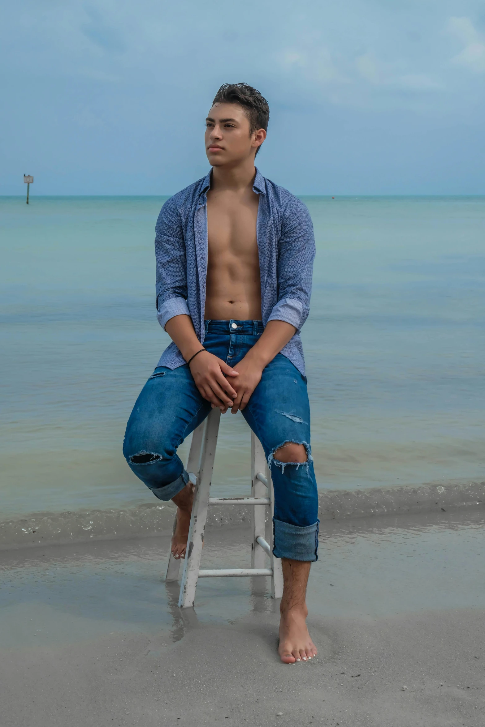 a man sitting on a stool on the beach, inspired by Byron Galvez, pexels contest winner, renaissance, gorgeous young model, wearing denim, androgynous male, florida man