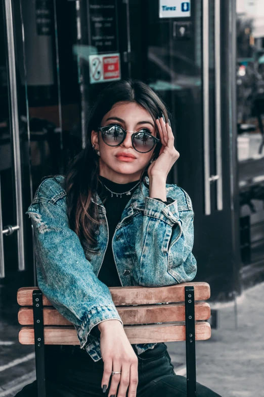a woman sitting on top of a wooden bench, trending on pexels, hurufiyya, wearing a jeans jackets, circular sunglasses, glam photo, teenager