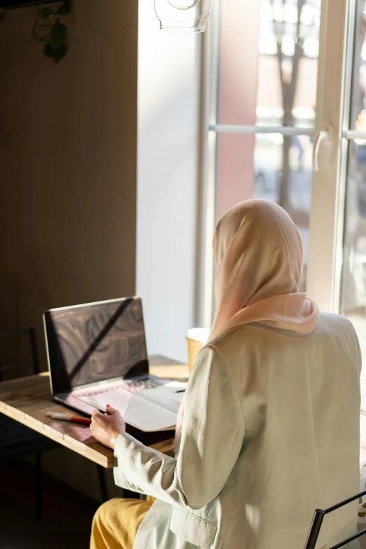 a woman sitting at a table in front of a laptop computer, a picture, inspired by Maryam Hashemi, trending on unsplash, hurufiyya, rear-shot, full frame image, veiled, sunlit