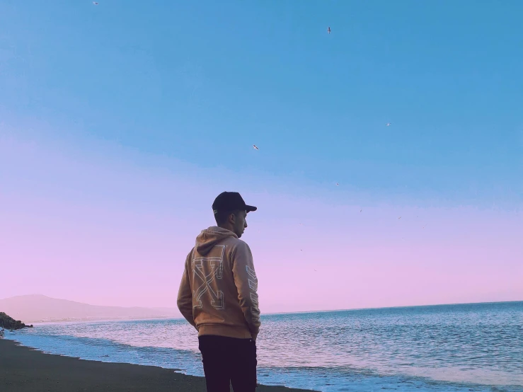 a man standing on a beach next to the ocean, an album cover, unsplash, realism, young man in a purple hoodie, the sky is pink, youtube thumbnail, background image
