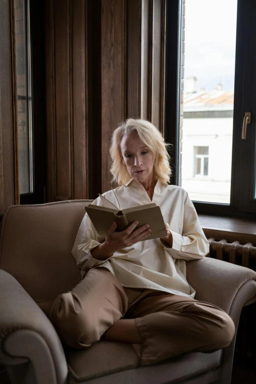 a woman sitting on a couch reading a book, a portrait, by Grytė Pintukaitė, trending on unsplash, elegant slim beige shirt, albino, aged, a blond