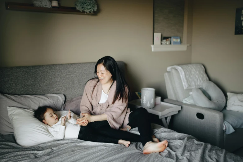 a woman sitting on top of a bed next to a baby, inspired by helen huang, pexels contest winner, happening, asian descent, manuka, grey, avatar image