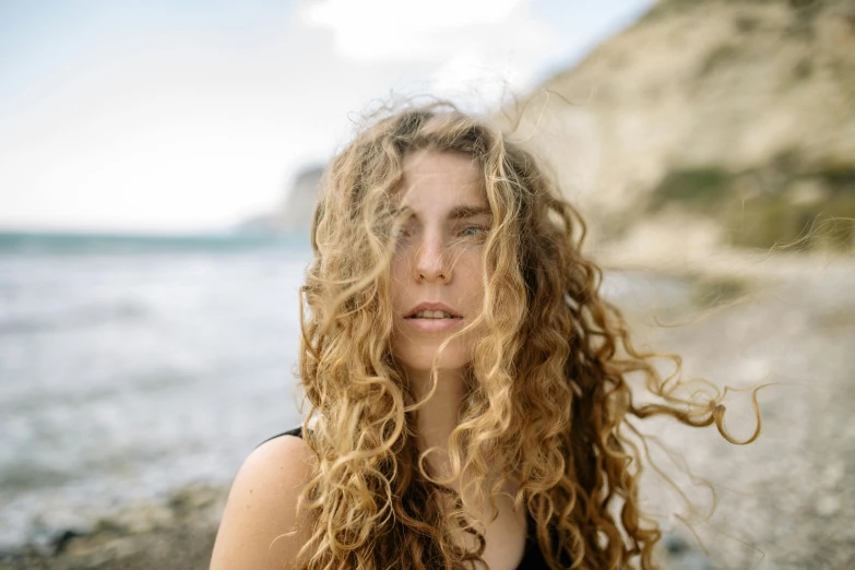 a woman with long curly hair standing in front of a body of water, trending on pexels, renaissance, bedhead, rugged face, portait image, near the beach