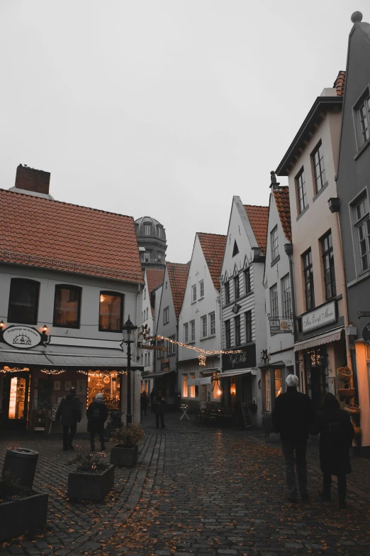 a group of people walking down a cobblestone street, by Christen Dalsgaard, pexels contest winner, art nouveau, white buildings with red roofs, overcast dusk, lower saxony, winter