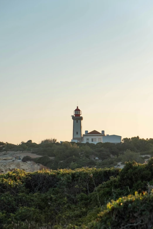 a lighthouse sitting on top of a lush green hillside, in a mediterranean lanscape, setting sun, square, farol da barra