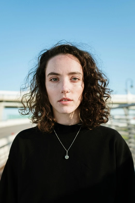 a woman with curly hair standing in front of a bridge, an album cover, unsplash contest winner, portrait androgynous girl, hyperrealism photo, demna gvasalia, wearing a black shirt