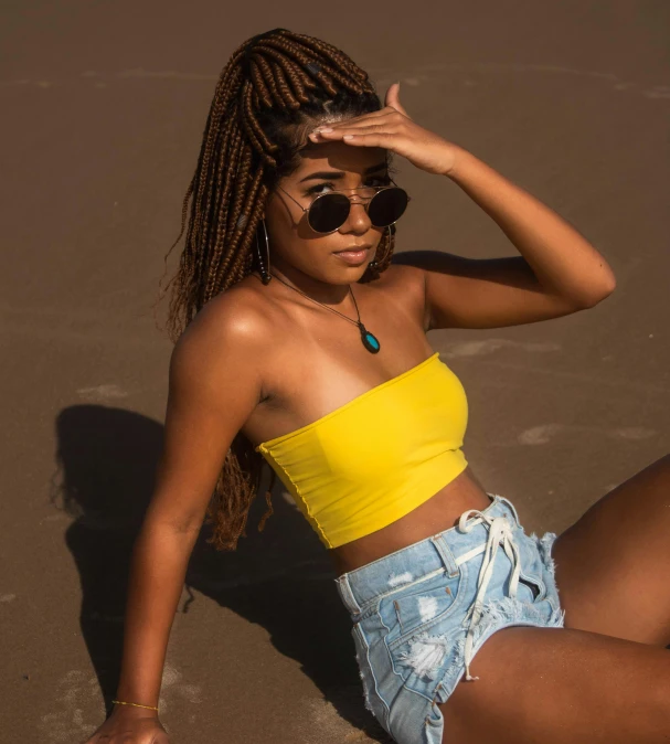 a woman sitting on top of a sandy beach, by Lily Delissa Joseph, trending on pexels, afrofuturism, wearing yellow croptop, dark shades, wearing denim short shorts, tubes