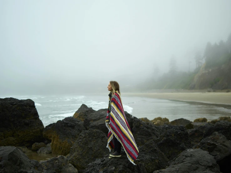 a woman standing on a rocky beach covered in a blanket, by Jessie Algie, foggy rainy day, press shot, sydney sweeney, looking into the distance