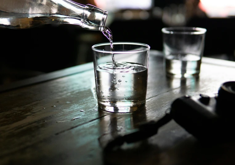 a person pouring water into a glass on a table, unsplash, purism, bottle of vodka, multiple stories, laura watson, on wooden table