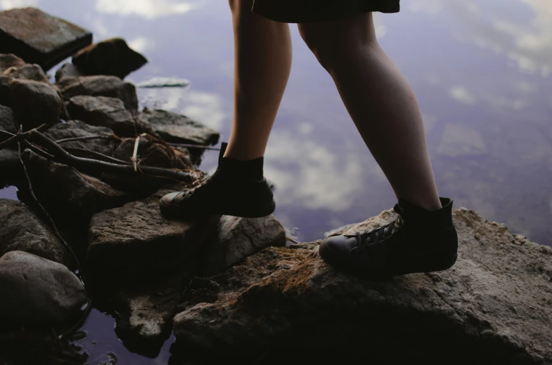 a person standing on a rock near a body of water, wearing black boots, side view of her taking steps, carson ellis, rocky roads