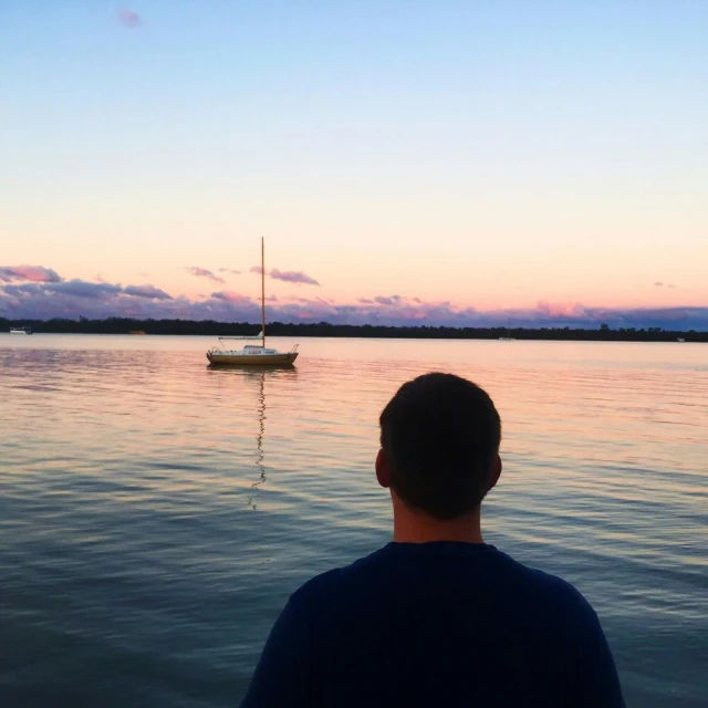 a man standing in front of a body of water with a sailboat in the distance, a picture, by Ryan Pancoast, happening, evening time, lachlan bailey, in a quiet moment, upon a peak in darien