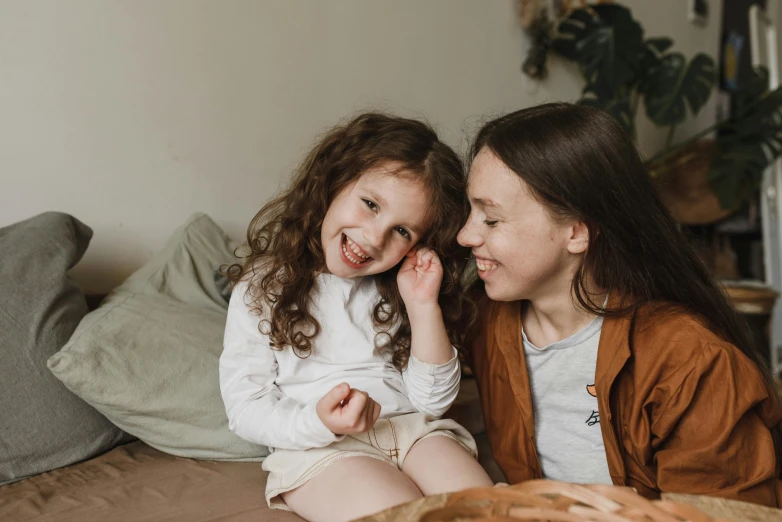 a couple of young girls sitting on top of a bed, by Emma Andijewska, pexels contest winner, she is smiling, brown curly hair, motherly, animated