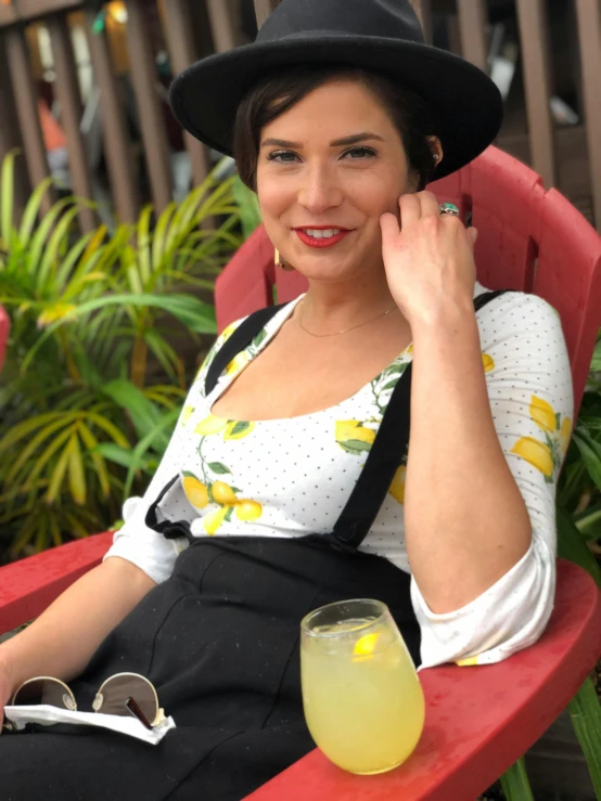 a woman sitting in a red chair with a drink, reddit, wearing a straw hat and overalls, wearing yellow floral blouse, wearing black camisole outfit, white straw flat brimmed hat