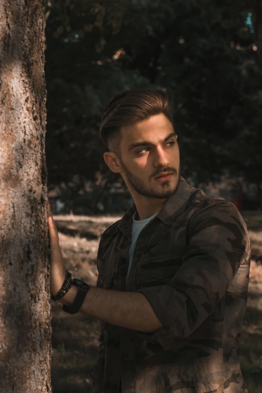 a man standing next to a tree in a forest, a colorized photo, by Adam Szentpétery, pexels contest winner, handsome attractive face, wearing camo, young greek man, lgbtq