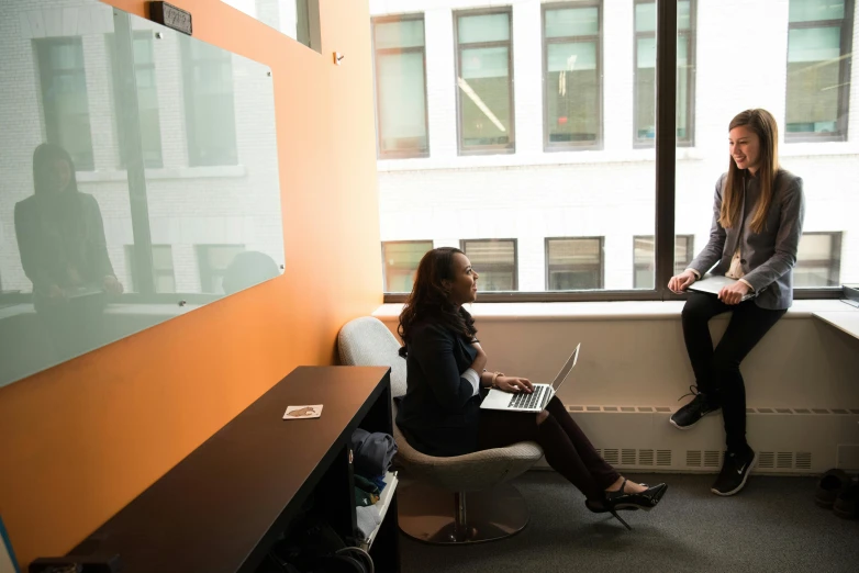 a couple of women sitting next to each other in a room, by Gavin Hamilton, unsplash, hurufiyya, cubical meeting room office, gray and orange colours, man sitting facing away, next to a window