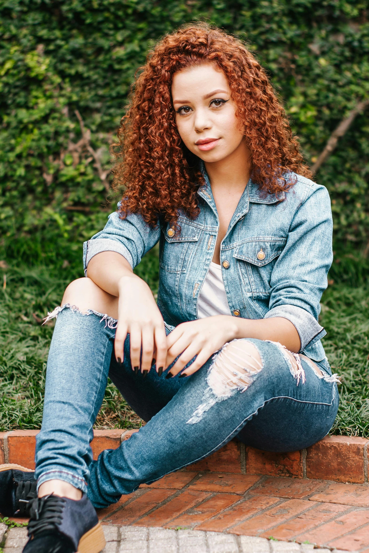 a beautiful young woman sitting on a brick wall, a portrait, by Leo Michelson, pexels, renaissance, wearing a jeans jackets, curly red hair, teenage vanessa morgan, promotional image