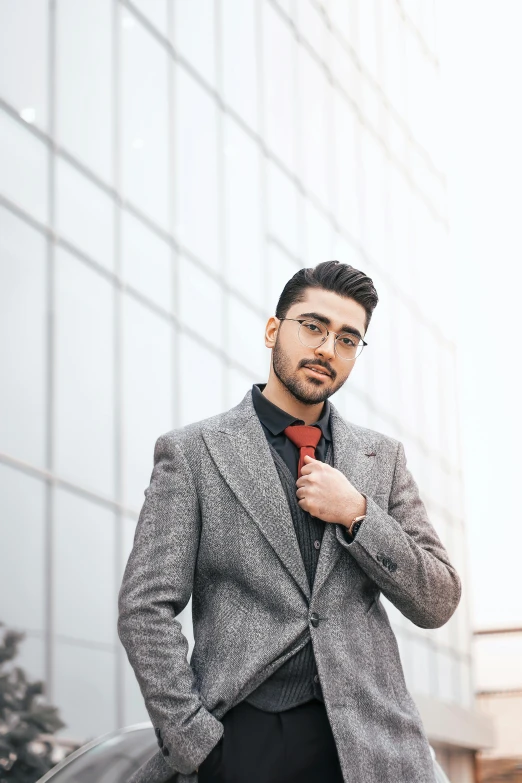 a man in a suit and tie standing in front of a building, by Ismail Acar, trending on pexels, renaissance, zayn malik, grey jacket, man with glasses, red sweater and gray pants