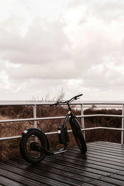 a motorcycle parked on top of a wooden deck, grey sky, electric, near the beach, black sokkel