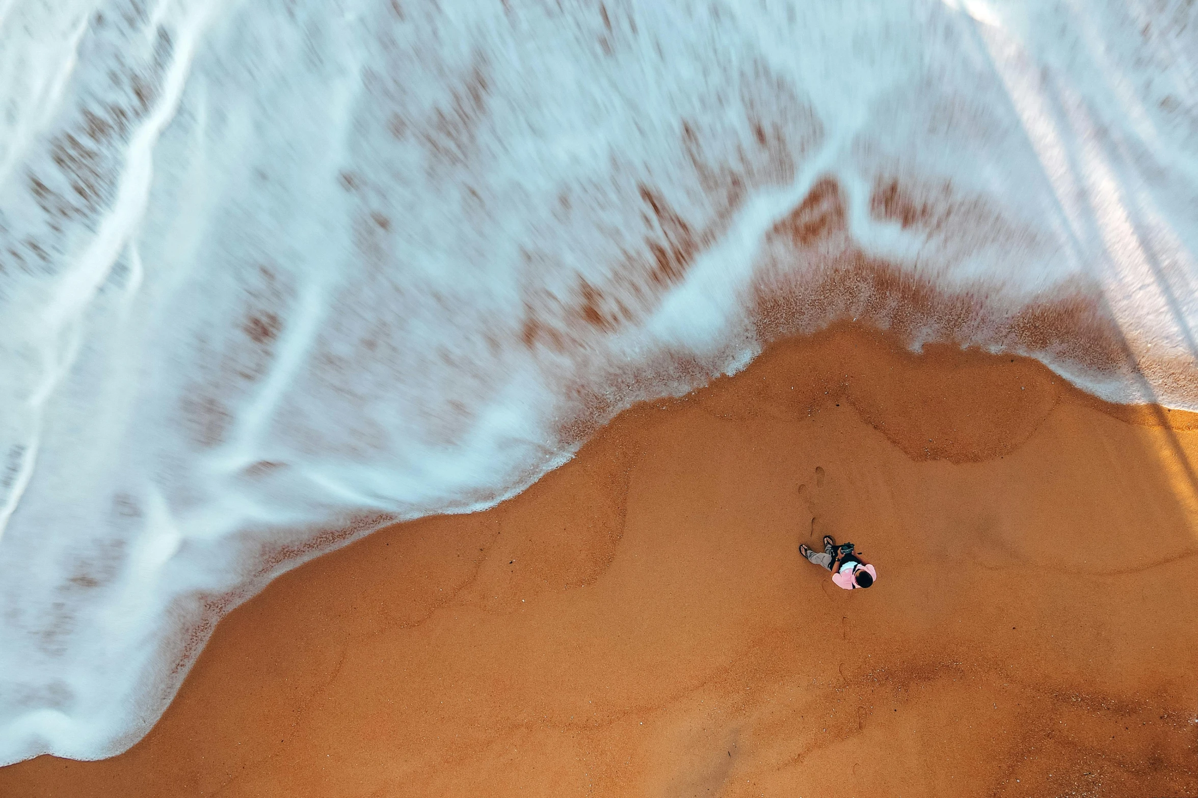 a person laying on top of a sandy beach next to the ocean, by Daniel Lieske, pexels contest winner, helicopter view, ocean swells, red sand, hd footage