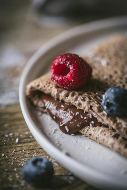 a white plate topped with a crepe covered in chocolate and raspberries, unsplash, bread, brown red blue, rustic, gif