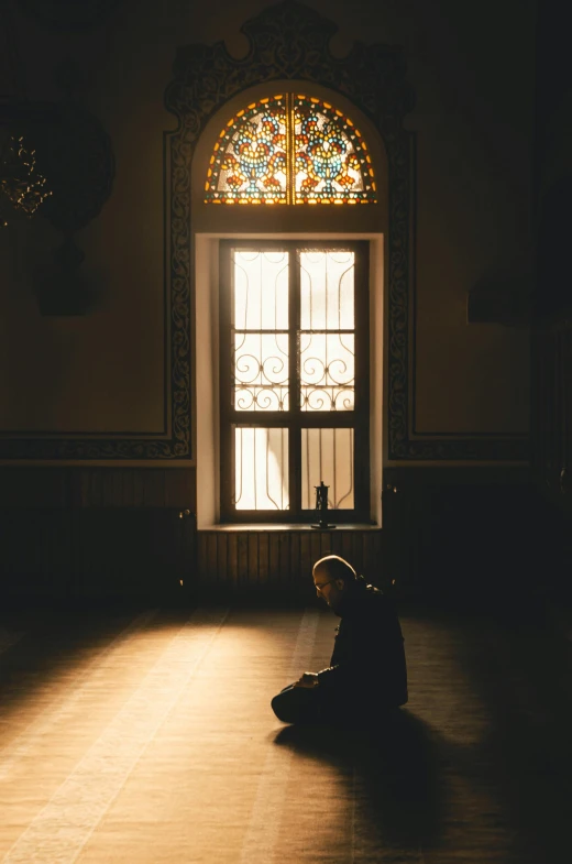 a person sitting on the floor in front of a window, by Matija Jama, pexels contest winner, hurufiyya, prayer, single light, highly ornate, hooded