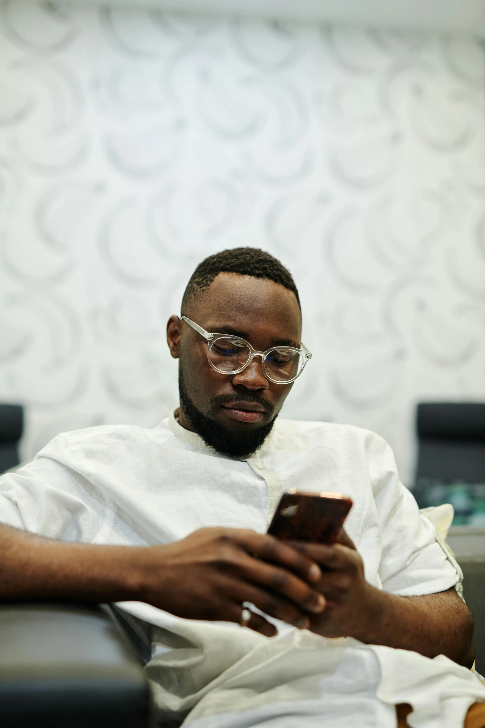 a man sitting in a chair looking at a cell phone, by Chinwe Chukwuogo-Roy, trending on pexels, in square-rimmed glasses, man with beard, jaylen brown, he is a long boi ”