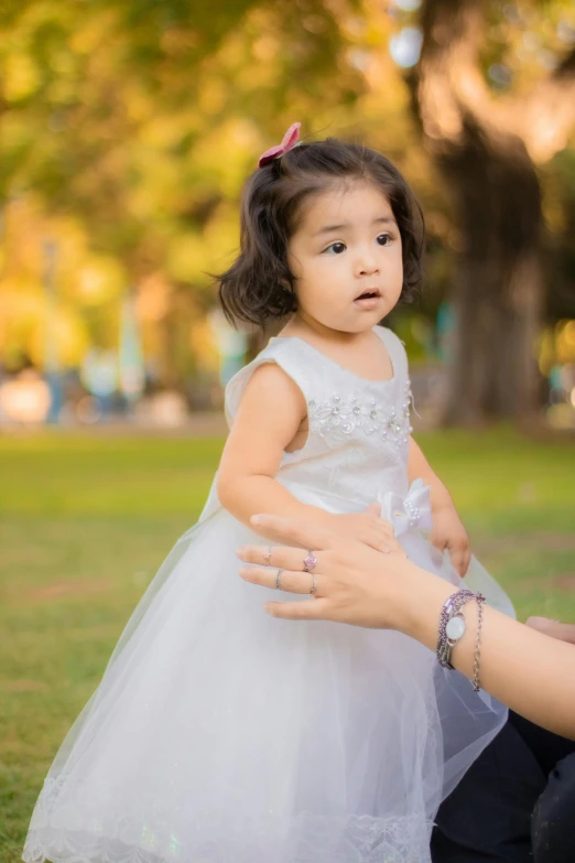 a woman holding a little girl in a white dress, by Lilia Alvarado, pexels contest winner, at a park, young asian girl, holding hand, a handsome