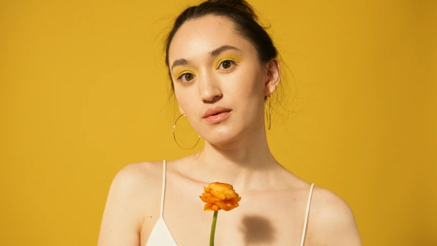 a woman holding a flower in front of her face, an album cover, inspired by Elsa Bleda, trending on pexels, photorealism, yellow-orange, portrait sophie mudd, gold eyeshadow, studio portrait photo