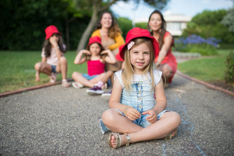 a little girl sitting on the ground in front of a group of girls, a picture, by Lilia Alvarado, pexels contest winner, red hat, avatar image, mini model, high resolution image