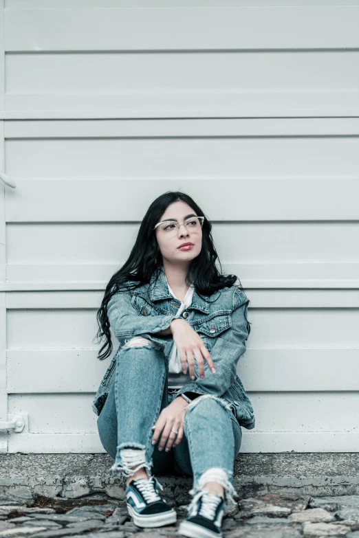 a woman sitting on the ground in front of a wall, pexels contest winner, antipodeans, wavy long black hair and glasses, wearing a jeans jackets, in front of white back drop, pouty