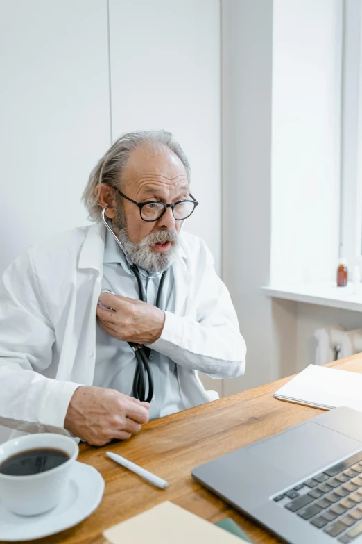 a man sitting at a desk with a laptop and a cup of coffee, wearing lab coat and glasses, coughing, grey beard, stethoscope