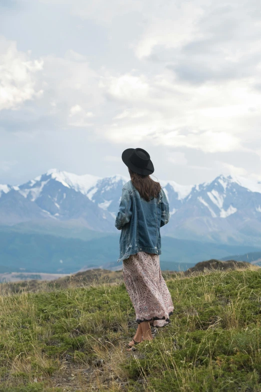 a woman standing on top of a lush green hillside, by Jessie Algie, trending on unsplash, snow capped mountains, alaska, movie still 8 k, back view also