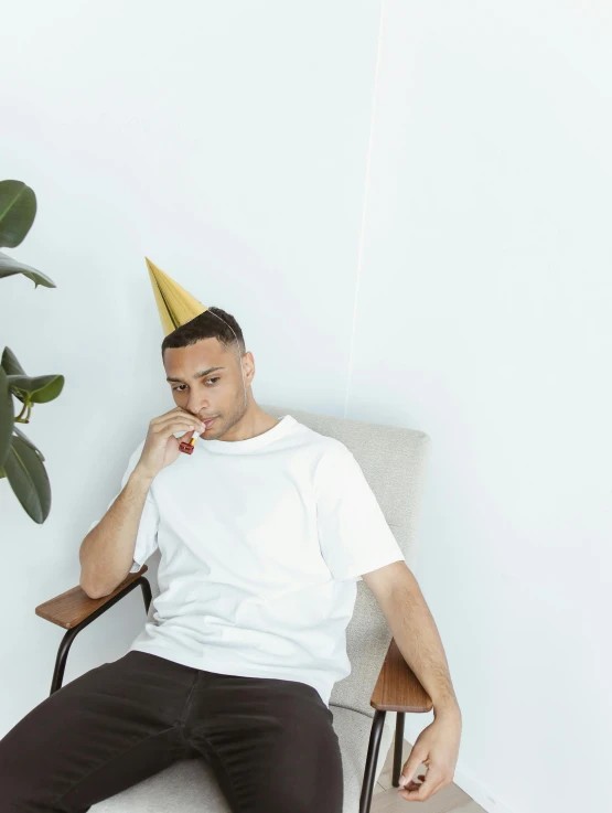 a man sitting in a chair wearing a party hat, by Robbie Trevino, white t-shirt, minimalist photo, gold shirt, tapered hairline