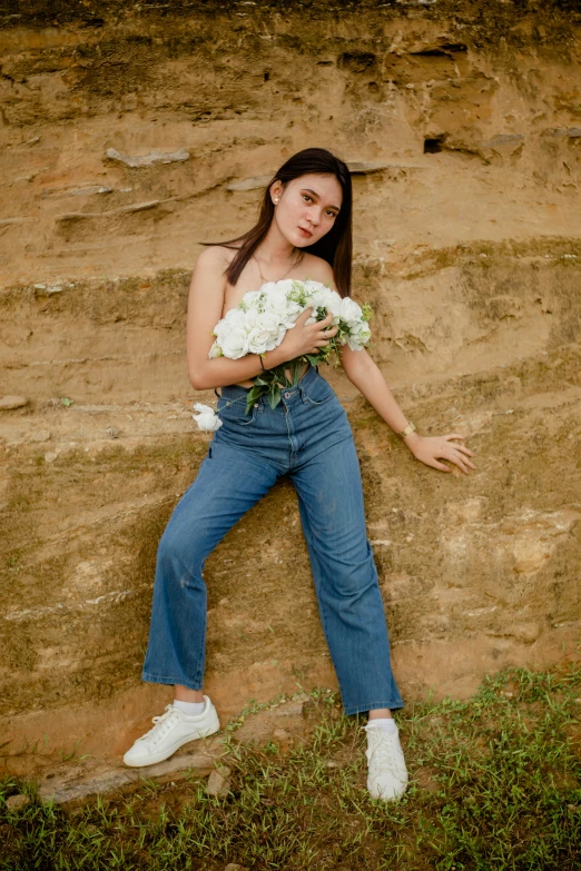 a woman leaning against a wall holding a bouquet of flowers, trending on pexels, wearing denim, in style of lam manh, on a rock, wearing a crop top