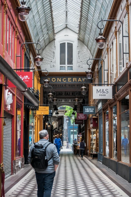 a man walking down a hallway in a building, quirky shops, northern france, payne's grey and venetian red, a busy arcade